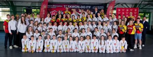 Zdjęcie przedstawia dużą grupę uczestników zawodów w strojach do taekwon-do na sali gimnastycznej. W tle plakat wydarzenia.